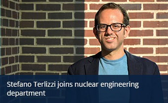 A person wearing glasses stands in front of a brick wall outside and smiles at the camera